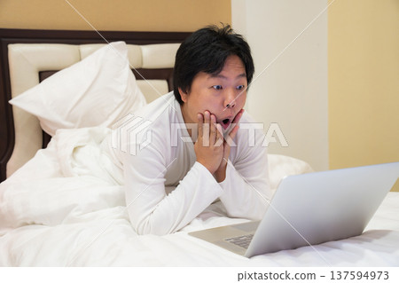 A man lies on his bed watching his laptop with a shocked expression. The image captures real emotion and surprise from something seen online. A man lies on his bed watching his laptop with a shocked expression. The image captures real emotion and surprise from something seen online. 137594973
