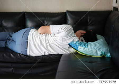 A man relaxes on the couch lying on his side facing away, taking a moment to rest in a calm home space that reflects stress relief and mental recharge. 137594976