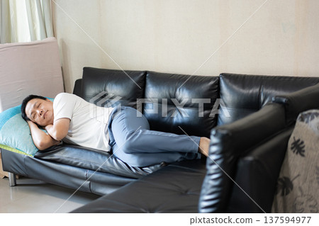 A man sleeps quietly on a black leather sofa in a cozy room. The calm atmosphere conveys rest, comfort, and everyday peace at home. 137594977