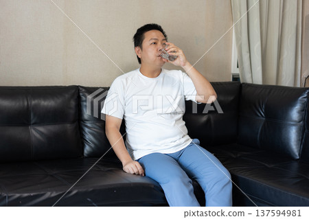A man drinks a glass of water while sitting on a black leather sofa, representing daily hydration and a healthy lifestyle. A man drinks a glass of water while sitting on a black leather sofa, representing daily hydration and a healthy lifestyle. 137594981