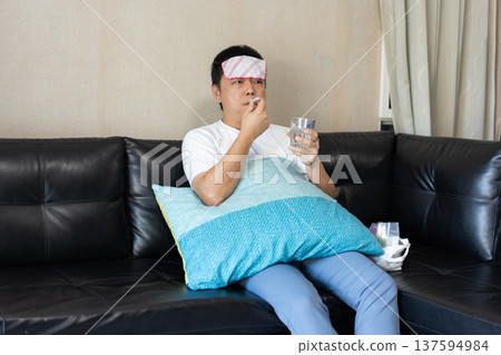 A man sits on a sofa with a wet towel on his forehead, holding a glass of water and medicine. The image shows illness and home recovery. A man sits on a sofa with a wet towel on his forehead, holding a glass of water and medicine. The image shows illness and home recovery. 137594984