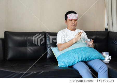 A man sits on a sofa with a wet towel on his forehead, holding a glass of water and medicine. The image shows illness and home recovery. 137594985