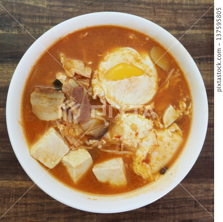 A bowl of spicy Korean tofu soup featuring soft tofu cubes, pork slices, kimchi, and a poached egg in rich red chili broth, a classic comforting dish known as Soondubu-jjigae at Asian restaurant 137595805