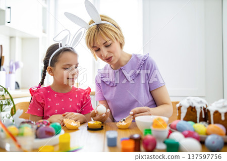 A mother and daughter wearing bunny ears are happily decorating Easter eggs with wax and paint at a wooden table 137597756