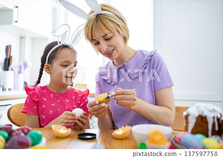 A mother and daughter wearing bunny ears are happily decorating Easter eggs with wax and paint at a wooden table A mother and daughter wearing bunny ears are happily decorating Easter eggs with wax and paint at a wooden table 137597758