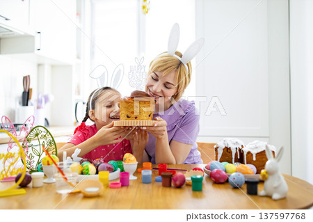 A mother and daughter wearing bunny ears excitedly sniff a traditional Easter cake while surrounded by painted eggs and baking supplies A mother and daughter wearing bunny ears excitedly sniff a traditional Easter cake while surrounded by painted eggs and baking supplies 137597768