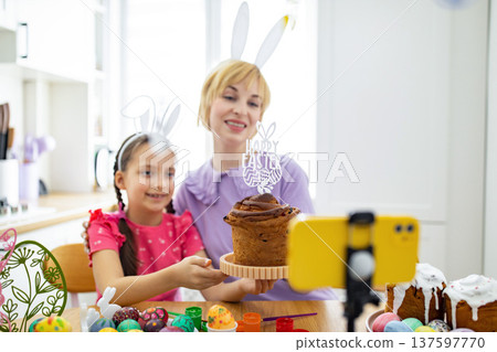 A mother and daughter wearing bunny ears are live streaming their Easter baking activities on a smartphone, surrounded by decorated eggs and cakes A mother and daughter wearing bunny ears are live streaming their Easter baking activities on a smartphone, surrounded by decorated eggs and cakes 137597770