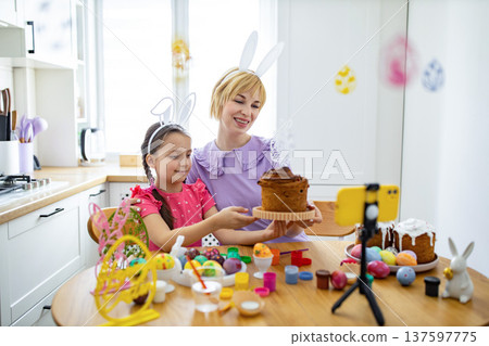 A mother and daughter wearing bunny ears are filming themselves baking and decorating Easter cakes and eggs in a bright kitchen A mother and daughter wearing bunny ears are filming themselves baking and decorating Easter cakes and eggs in a bright kitchen 137597775
