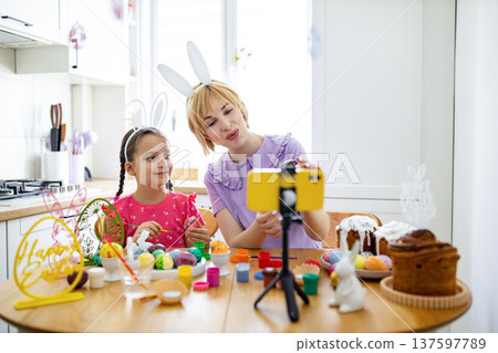 A mother and daughter wearing bunny ears are decorating Easter eggs and filming their activity on a smartphone A mother and daughter wearing bunny ears are decorating Easter eggs and filming their activity on a smartphone 137597789