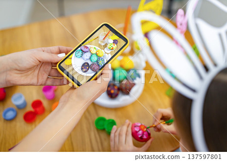 A person captures a photo of beautifully decorated Easter eggs on their smartphone while another person paints an egg, surrounded by craft supplies A person captures a photo of beautifully decorated Easter eggs on their smartphone while another person paints an egg, surrounded by craft supplies 137597801