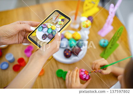 A mother and child are painting and decorating Easter eggs together on a wooden table, capturing the moment with a smartphone 137597802