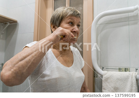 Senior woman actively brushing her teeth in the bathroom, maintaining good oral hygiene as part of her daily self-care routine. The real life of an elderly person, daily routine Senior woman actively brushing her teeth in the bathroom, maintaining good oral hygiene as part of her daily self-care routine. The real life of an elderly person, daily routine 137599195