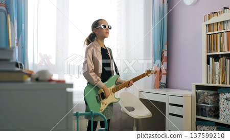 Teen girl in sunglasses plays electric guitar at home, exploring her passion and enjoying the creative process in a colorful room 137599210