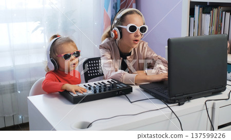 Two girls enjoy making music at home together. Older sister plays midi keyboard while younger follows along, focused on their craft Two girls enjoy making music at home together. Older sister plays midi keyboard while younger follows along, focused on their craft 137599226