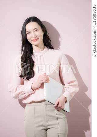 Cute Japanese woman in office casual wear holding a laptop on a pink background, smiling and looking at the camera 137599539