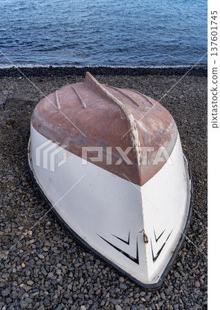 An old, overturned rowboat rests on a pebble beach, facing the viewer with the ocean visible in the background on a cloudy day. An old, overturned rowboat rests on a pebble beach, facing the viewer with the ocean visible in the background on a cloudy day. 137601745