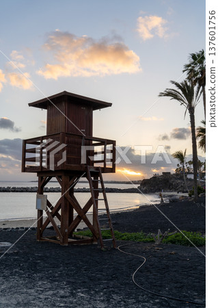 A wooden lifeguard tower stands on a black sand beach in Lanzarote, overlooking the sea as the sun sets in the background. 137601756