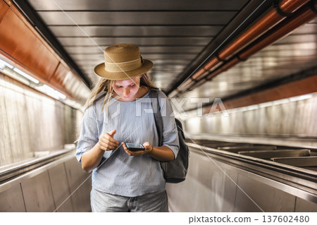 Young woman using smartphone while descending escalator in metro tunnel, urban travel lifestyle Young woman using smartphone while descending escalator in metro tunnel, urban travel lifestyle 137602480