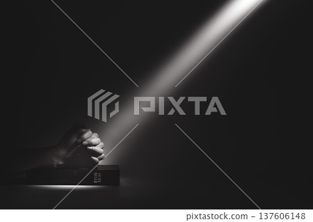 A strong beam of light illuminating a holy bible and the hands of a person praying, a background of faith and belief. A strong beam of light illuminating a holy bible and the hands of a person praying, a background of faith and belief. 137606148