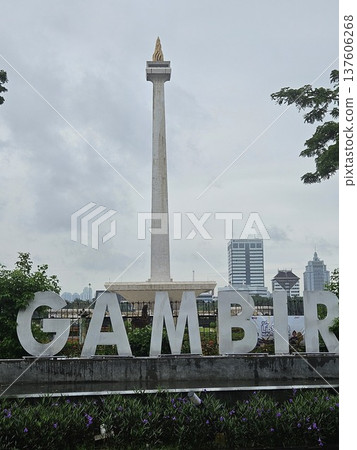 The National Monument of Indonesia stands tall behind a large white Gambir sign under a cloudy sky in Jakarta, surrounded by lush green trees and modern city buildings in a scenic urban landscape. The National Monument of Indonesia stands tall behind a large white Gambir sign under a cloudy sky in Jakarta, surrounded by lush green trees and modern city buildings in a scenic urban landscape. 137606268