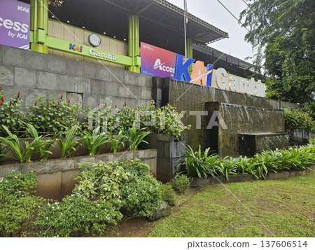 The iconic facade of Gambir Railway Station in Jakarta features the bold KAI Gambir signage above a tiered stone water fountain, surrounded by lush green garden plants under a gloomy overcast sky. The iconic facade of Gambir Railway Station in Jakarta features the bold KAI Gambir signage above a tiered stone water fountain, surrounded by lush green garden plants under a gloomy overcast sky. 137606514