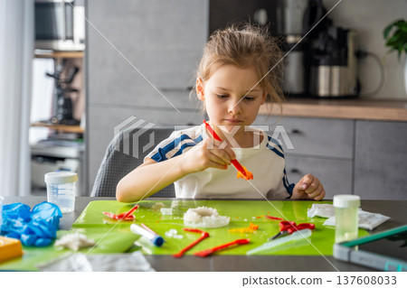 Young girl carefully holding grown crystal with tweezers and examining it during home science experiment. Precision work and analytical thinking in early STEM education and chemistry learning. 137608033