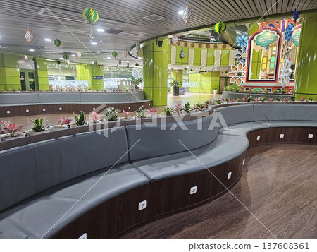 A modern waiting lounge featuring curved grey cushioned benches with integrated power outlets, decorated with festive green hanging ornaments and potted plants under a contemporary slatted ceiling. 137608361