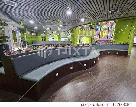A modern waiting lounge featuring curved grey cushioned benches with integrated power outlets, decorated with festive green hanging ornaments and potted plants under a contemporary slatted ceiling. A modern waiting lounge featuring curved grey cushioned benches with integrated power outlets, decorated with festive green hanging ornaments and potted plants under a contemporary slatted ceiling. 137608363