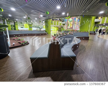 A modern waiting lounge featuring curved grey cushioned benches with integrated power outlets, decorated with festive green hanging ornaments and potted plants under a contemporary slatted ceiling. 137608367
