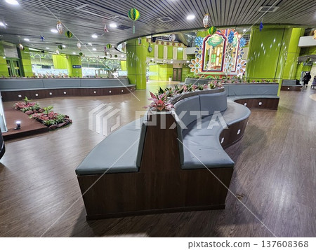 A modern waiting lounge featuring curved grey cushioned benches with integrated power outlets, decorated with festive green hanging ornaments and potted plants under a contemporary slatted ceiling. A modern waiting lounge featuring curved grey cushioned benches with integrated power outlets, decorated with festive green hanging ornaments and potted plants under a contemporary slatted ceiling. 137608368