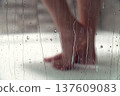 Female Bare feet of a person standing on a wet shower floor, with water droplets on a glass door creating a blurred effect in a modern bathroom setting 137609083