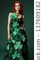Female model with red hair wearing a green dress adorned with shamrock shapes, standing against a dark green background, showcasing a festive Saint Patrick's Day theme 137609182