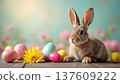 Brown rabbit sitting beside colorful Easter eggs and a yellow flower on a wooden surface, with a soft blurred background of pastel colors and floral elements 137609222
