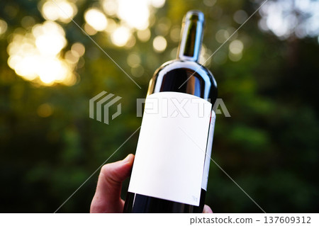 Red wine bottle with blank white label with space for text. Wine bottle in hand on green leaves Red wine bottle with blank white label with space for text. Wine bottle in hand on green leaves 137609312