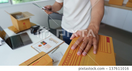 E-commerce Management. Young man organizing packages while checking inventory on tablet. E-commerce Management. Young man organizing packages while checking inventory on tablet. 137611016