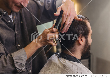 Barber cutting male client hair with scissors and comb. Close up of professional haircut process, precision technique, men grooming service in modern barbershop. Barber cutting male client hair with scissors and comb. Close up of professional haircut process, precision technique, men grooming service in modern barbershop. 137611492