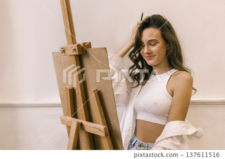 Young female artist standing near easel and holding pencil while working on painting in art studio, creative process and inspiration 137611516
