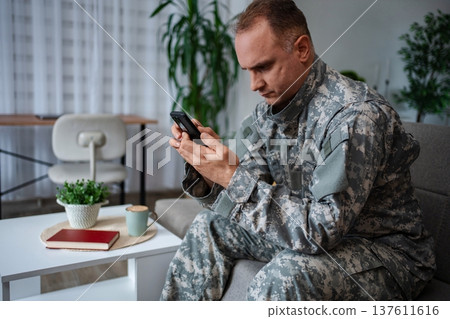Soldier sitting on sofa using smartphone at home Soldier sitting on sofa using smartphone at home 137611616