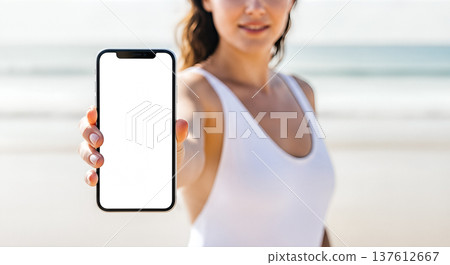Beautiful young woman wearing white swimsuit showing blank smartphone screen ocean beach background, summer vacation travel mobile app advertising presentation mockup concept 137612667