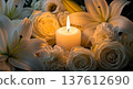 Glowing burning white candle standing among beautiful fresh white roses and lilies flowers creating warm romantic peaceful memorial atmosphere dark background elegant composition 137612690