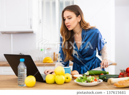 Young woman in blue robe cooking and watching TV series on laptop 137615681