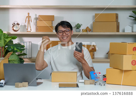 E-commerce Success. Young entrepreneur celebrating online sales success with smartphone in home office. E-commerce Success. Young entrepreneur celebrating online sales success with smartphone in home office. 137616888