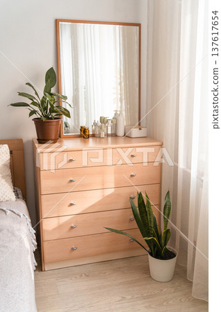 Wooden dresser in a modern bedroom interior, displaying a mirror, decorative plant, and various beauty bottles on top 137617654
