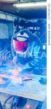 Vertical. A welder wearing a protective mask performs arc welding of metal parts, generating bright sparks and an intense glow in an industrial workshop.. Vertical. A welder wearing a protective mask performs arc welding of metal parts, generating bright sparks and an intense glow in an industrial workshop.. 137620132