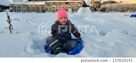 A little girl sits on a blue sled in freshly fallen snow, wearing a pink hat and warm winter clothes. The child enjoys playing outdoors on a cold winter day. The child slides down an icy hill. A little girl sits on a blue sled in freshly fallen snow, wearing a pink hat and warm winter clothes. The child enjoys playing outdoors on a cold winter day. The child slides down an icy hill. 137620141