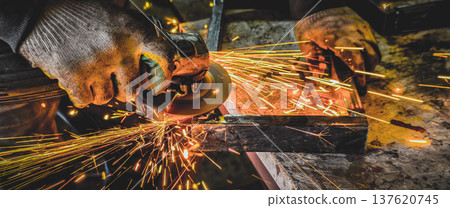 Worker hands wearing protective gloves operating an angle grinder, creating a shower of bright orange sparks while cutting or shaping metal in an industrial workshop. Worker hands wearing protective gloves operating an angle grinder, creating a shower of bright orange sparks while cutting or shaping metal in an industrial workshop. 137620745