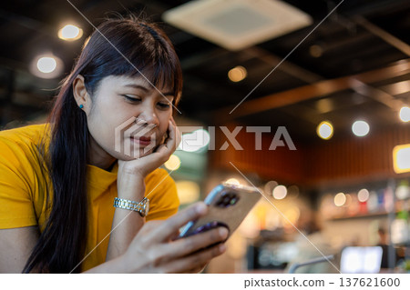 Asian woman in yellow t-shirt using smartphone while sitting in cafe. Concept of mobile communication, social media browsing, reading news, and digital lifestyle in modern coffee shop. Asian woman in yellow t-shirt using smartphone while sitting in cafe. Concept of mobile communication, social media browsing, reading news, and digital lifestyle in modern coffee shop. 137621600