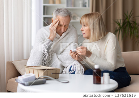 Senior woman checking thermometer while caring for sick husband wrapped in blanket 137621619