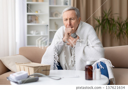 Senior man sitting on sofa wrapped in blanket while coughing at home 137621634