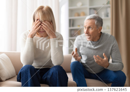 Senior woman covering her face with hands while her husband speaks emotionally Senior woman covering her face with hands while her husband speaks emotionally 137621645